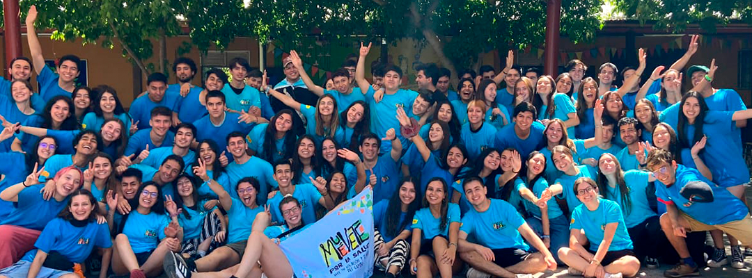 Grupo de más de 60 estudiantes de Medicina UC posa para la fotografía sentados en el suelo, en un lugar exterior, bajo un toldo. Todos sonríen y muchos levantan sus manos. Todos llevan puesta una polera celeste del programa de voluntariado Muévete por la Salud. Al centro, dos jóvenes estudiantes sostienen una bandera donde se lee "Muévete".