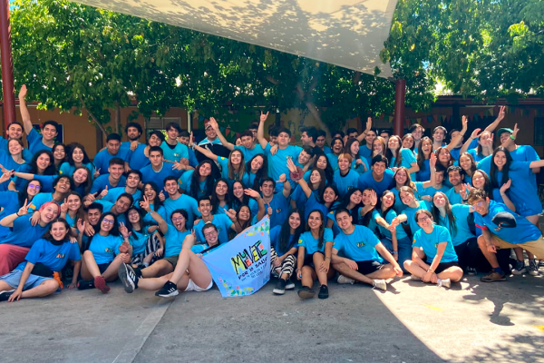 Grupo de más de 60 estudiantes de Medicina UC posa para la fotografía sentados en el suelo, en un lugar exterior, bajo un toldo. Todos sonríen y muchos levantan sus manos. Todos llevan puesta una polera celeste del programa de voluntariado Muévete por la Salud. Al centro, dos jóvenes estudiantes sostienen una bandera donde se lee "Muévete".